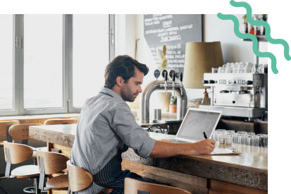 A male restaurant worker sitting at a counter writing something down as they look at their computer.